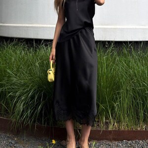 May include: A black midi-length dress with a V-neckline and short sleeves, featuring a delicate black lace trim along the hemline. The dress is paired with a small, bright yellow structured handbag and clear heeled sandals with black accents.
