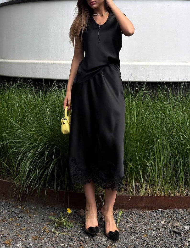 May include: A black midi-length dress with a V-neckline and short sleeves, featuring a delicate black lace trim along the hemline. The dress is paired with a small, bright yellow structured handbag and clear heeled sandals with black accents.
