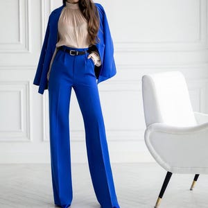 May include: A vibrant blue pantsuit, featuring wide-leg trousers and a matching blazer draped over the shoulders. The outfit includes a beige blouse and a black belt. The ensemble is set against a white backdrop, with a white chair in the background.