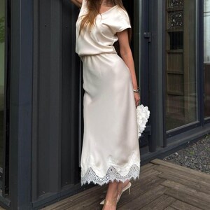 May include: A cream-colored satin midi dress featuring short sleeves, a round neckline, and a white lace trim along the hemline. The dress has a relaxed bodice and a straight skirt silhouette, paired with pointed-toe heels and a white floral clutch.