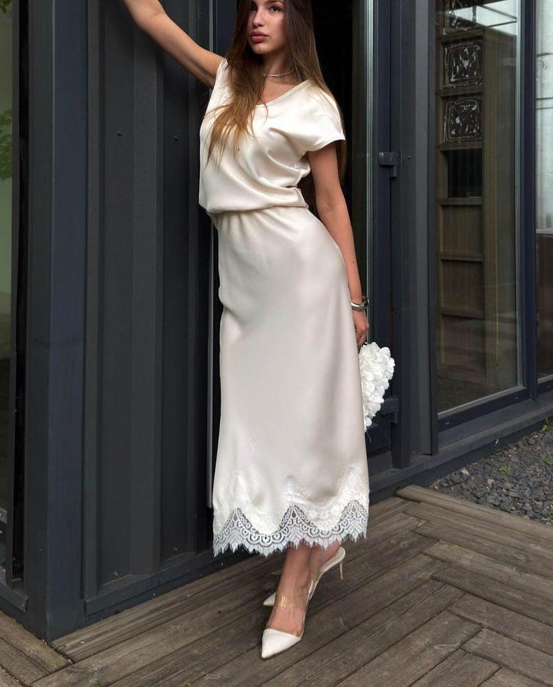 May include: A cream-colored satin midi dress featuring short sleeves, a round neckline, and a white lace trim along the hemline. The dress has a relaxed bodice and a straight skirt silhouette, paired with pointed-toe heels and a white floral clutch.