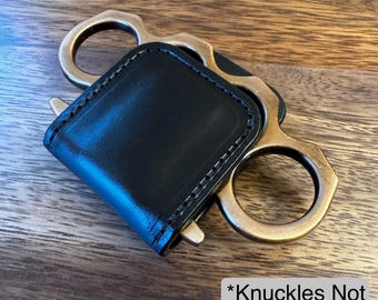 May include: A black leather cover with visible stitching designed to fit over a brass-colored metal knuckle duster. The item features a rectangular leather piece folded over the metal frame. A white label with black text reads *Knuckles Not Included*.