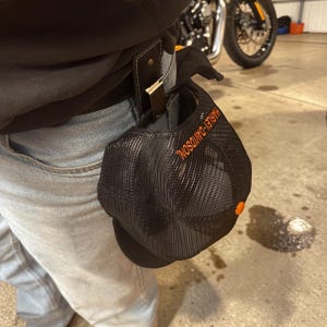 Puede incluir: Gorra de béisbol Harley-Davidson de malla negra con letras naranjas y un pequeño botón naranja. La gorra está sujeta a una trabilla de cinturón de cuero negro.