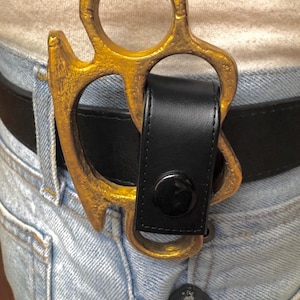May include: A gold-coloured belt buckle shaped like brass knuckles, attached to a black leather belt. The buckle is worn over light blue denim jeans. A black leather loop with a button secures the buckle.