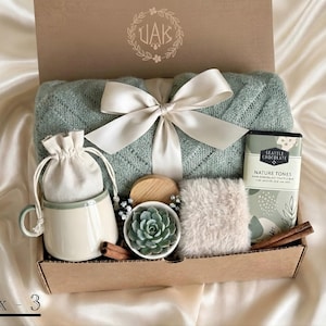 Könnte beinhalten: Geschenkbox mit einer hellgrünen Decke, die mit einer weißen Schleife gebunden ist. Enthält eine Tasse, einen kleinen Beutel, eine Sukkulente, eine Schokoladentafel, ein flauschiges Quadrat und Zimtstangen. Die Box ist mit "Box - 3" beschriftet.