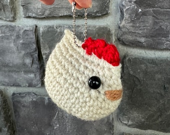 May include: A cream-colored crocheted chicken keychain featuring a bright red textured comb, a single black plastic bead eye, and a small tan beak. The item is attached to a silver-toned metal ball chain for hanging and display purposes.