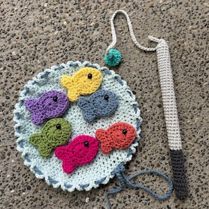 Puede incluir: Un juego de pesca de crochet con una base circular azul y blanca y seis peces de colores. Los peces son morado, amarillo, verde, rojo y rosa. Se incluye una caña de pescar de crochet gris con un flotador azul y verde.