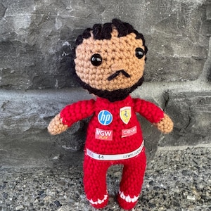 May include: A crocheted doll with dark hair and a beard, wearing a red racing suit. The suit features logos for HP, VGW Play, Ceva, and a Ferrari emblem. A white belt across the waist displays the text "44" and "LEWIS HAMILTON" with a small UK flag.