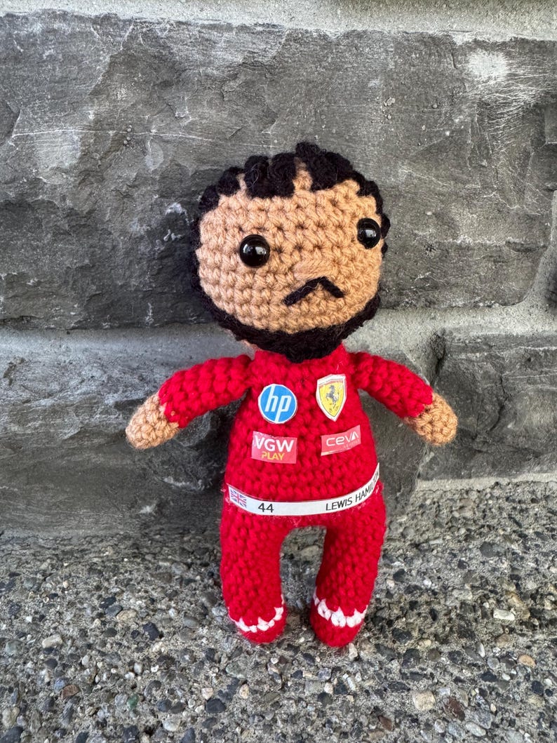 May include: A crocheted doll with dark hair and a beard, wearing a red racing suit. The suit features logos for HP, VGW Play, Ceva, and a Ferrari emblem. A white belt across the waist displays the text "44" and "LEWIS HAMILTON" with a small UK flag.