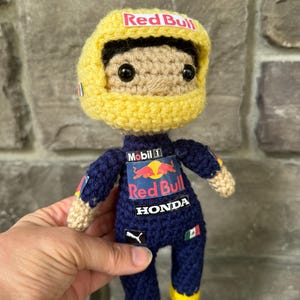 May include: A crocheted doll wearing a dark blue racing suit with Red Bull, Mobil 1, Honda, and Puma logos. The doll has a tan face, black eyes, and a yellow racing helmet with a Red Bull logo on the front and a small Mexican flag on the side.