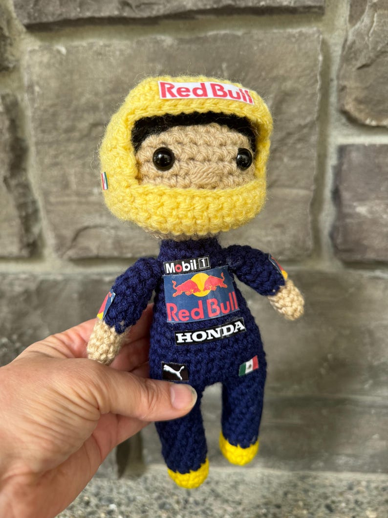 May include: A crocheted doll wearing a dark blue racing suit with Red Bull, Mobil 1, Honda, and Puma logos. The doll has a tan face, black eyes, and a yellow racing helmet with a Red Bull logo on the front and a small Mexican flag on the side.