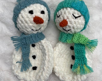 May include: Two crocheted snowman figures made of white yarn. The left snowman wears a blue hat and scarf with black eyes and buttons. The right snowman has a green and teal hat and scarf, closed eyes, and black buttons. Both have orange noses.