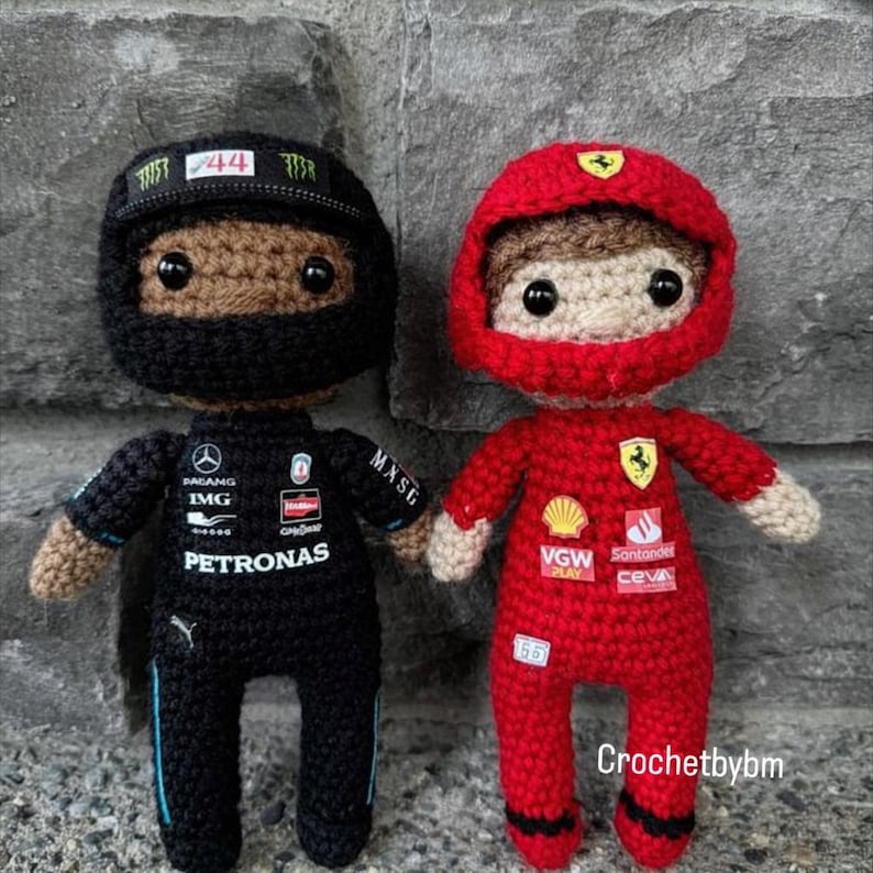 May include: Two crochet dolls representing Formula 1 drivers. The left doll wears a black racing suit with Petronas branding and a black helmet with the number 44. The right doll wears a red racing suit with Ferrari branding and a red helmet.