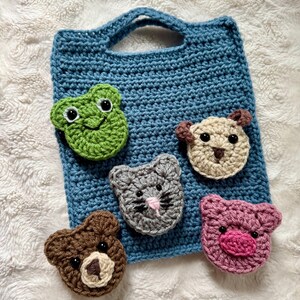 May include: A blue crocheted bag with a handle, featuring five crocheted animal faces: a frog, a dog, a cat, a bear, and a pig. The animals are attached to the bag and have embroidered details.
