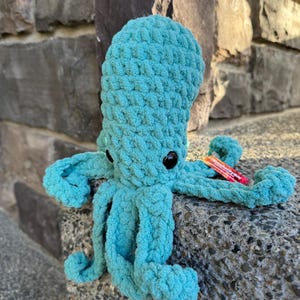 May include: A turquoise crocheted octopus plush toy with black button eyes. The handmade sea creature has eight tentacles and is sitting on a stone surface. The plush toy is made of a soft, textured yarn.