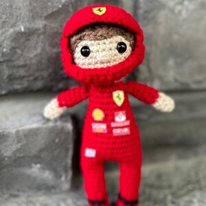 May include: A crocheted doll dressed in a red racing suit and matching helmet. The suit features small patches with logos including a yellow shield, a red and white rectangular emblem, and text. The doll has brown hair, black eyes, and light skin.