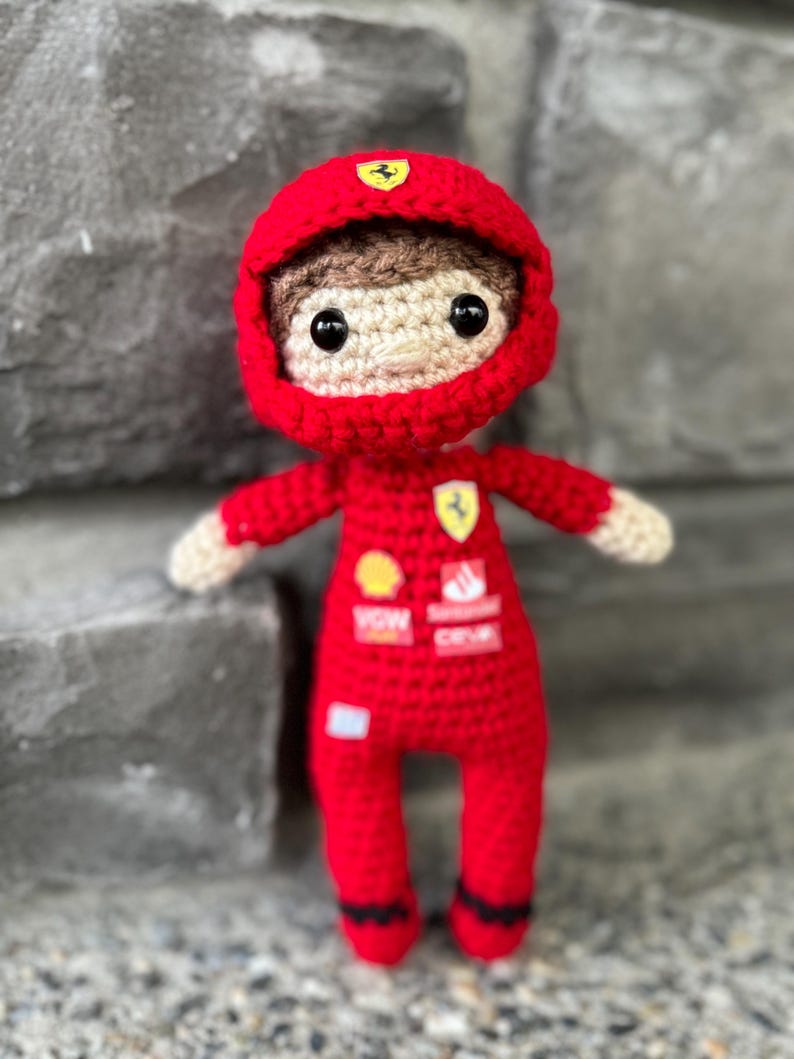 May include: A crocheted doll dressed in a red racing suit and matching helmet. The suit features small patches with logos including a yellow shield, a red and white rectangular emblem, and text. The doll has brown hair, black eyes, and light skin.