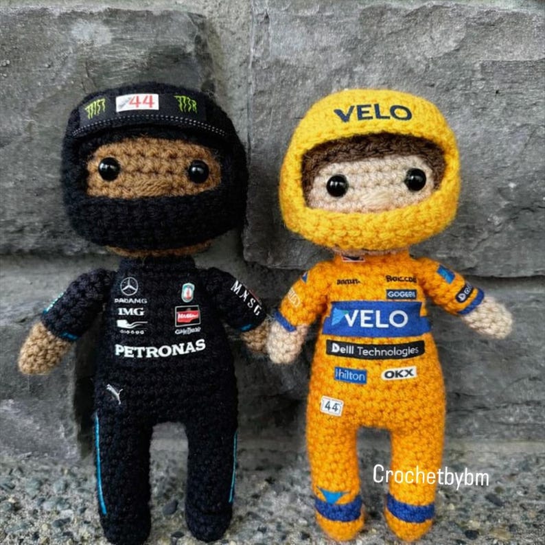 May include: Two crochet dolls representing race car drivers. The left doll wears a black racing suit with Petronas branding and a black helmet with the number 44. The right doll wears an orange racing suit with Velo branding and a yellow helmet.