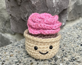 Peut inclure: Une petite plante succulente crochetée à la main dans un pot beige avec un visage souriant noir brodé. La plante présente des pétales en laine rose vif superposés sur une base marron foncé, formant un objet décoratif en fibre.