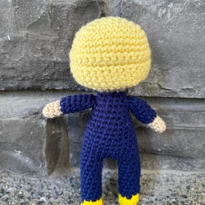 May include: A handmade crochet doll featuring a large, round yellow head, a dark blue long-sleeved jumpsuit, and bright yellow feet. The doll has light beige hands extending from the dark blue sleeves and is constructed with visible yarn stitches.