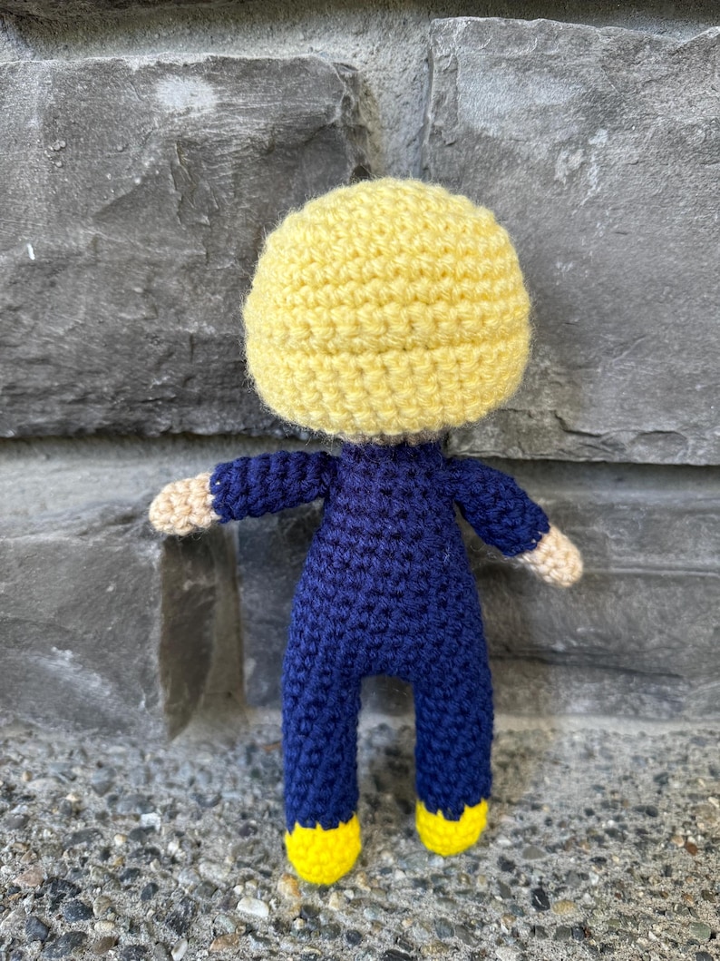 May include: A handmade crochet doll featuring a large, round yellow head, a dark blue long-sleeved jumpsuit, and bright yellow feet. The doll has light beige hands extending from the dark blue sleeves and is constructed with visible yarn stitches.