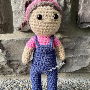 May include: A crocheted doll wearing blue overalls, a pink shirt, and a brown and pink headband. The doll has brown yarn hair and black eyes. The doll is standing on a bed of small rocks. The text "@CrochetbyBM" is visible on the doll's overalls.