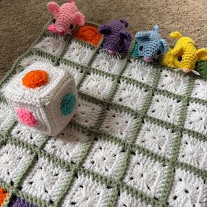 Könnte beinhalten: Eine handgefertigte Häkeldecke für Babys mit einem weißen und grünen Quadratmuster. Die Decke ist mit vier bunten Häkelmäusen und einem weißen Würfel mit bunten Punkten verziert. Die Mäuse sind rosa, lila, blau und gelb.
