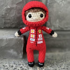 May include: A crocheted doll of a race car driver wearing a red racing suit with a hood. The suit features various sponsor logos including Shell, Ferrari, Ray-Ban, VGV, and Santander. The doll has brown hair, black eyes, and black cuffs.