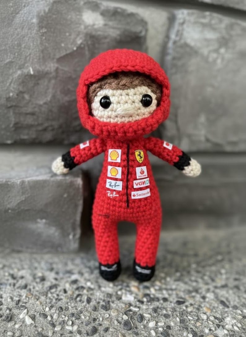 May include: A crocheted doll of a race car driver wearing a red racing suit with a hood. The suit features various sponsor logos including Shell, Ferrari, Ray-Ban, VGV, and Santander. The doll has brown hair, black eyes, and black cuffs.