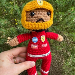 May include: A crocheted doll wearing a yellow helmet and a red racing suit. The suit features logos for HP, VGW Play, Ceva, and a Ferrari emblem. A white belt displays the number 44, a British flag, and the text Lewis Hamilton in black lettering.