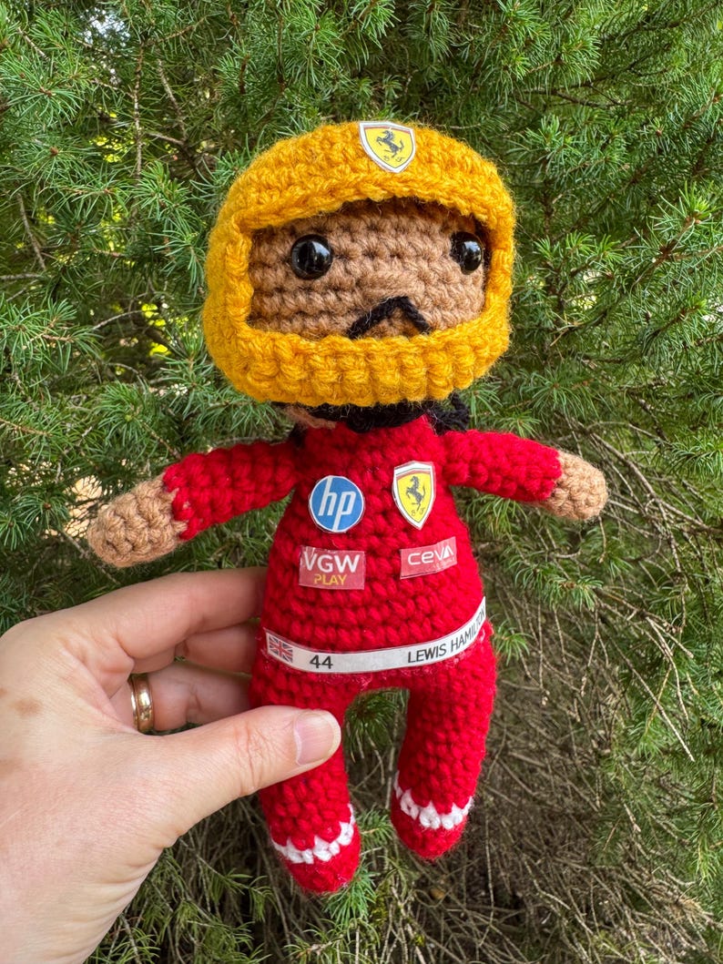 May include: A crocheted doll wearing a yellow helmet and a red racing suit. The suit features logos for HP, VGW Play, Ceva, and a Ferrari emblem. A white belt displays the number 44, a British flag, and the text Lewis Hamilton in black lettering.
