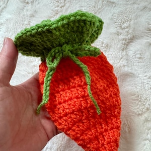 Puede incluir: Bolsa con forma de zanahoria tejida a ganchillo, naranja con la parte superior y el cordón verdes. El objeto se sostiene en una mano sobre un fondo blanco texturizado. El artículo probablemente se utiliza para guardar cosas o como objeto decorativo.