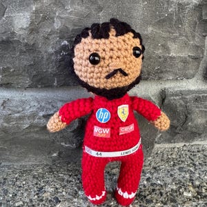 May include: A crocheted doll with dark hair and a beard, wearing a red racing suit. The suit features logos for HP, VGW Play, Ceva, and a Ferrari emblem. A white belt across the waist displays the text "44" and "LEWIS HAMILTON" with a small UK flag.