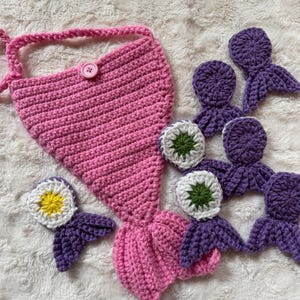May include: A pink crocheted mermaid tail with a single pink button at the top, accompanied by several smaller purple crocheted mermaid tail motifs. Some of these smaller purple motifs feature white and green or white and yellow circular centers.