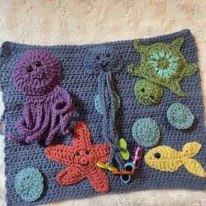 May include: A rectangular blue crochet mat featuring various sea creatures including a purple octopus, a green turtle, a coral starfish, and a yellow fish. The mat also includes small circular accents and a jellyfish with long dangling tentacles.