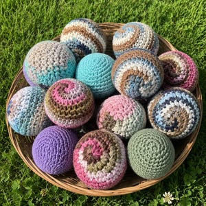 May include: A collection of round, crocheted yarn balls in a woven basket. The spheres feature various color combinations including shades of blue, brown, pink, purple, green, and white, with some displaying solid colors and others showing spiral patterns.