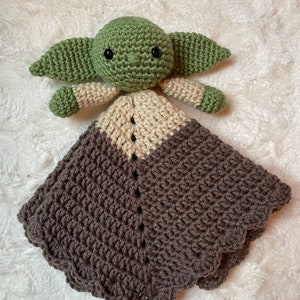 May include: A crocheted baby blanket with a green and beige character in the center. The character has large eyes and a small mouth. The blanket is brown with a scalloped edge.