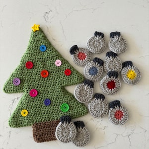 May include: A green crocheted Christmas tree with a brown trunk, decorated with a yellow star button and various colorful round buttons. Beside the tree are several small, circular, grey crocheted ornaments with black tops and colorful centers.