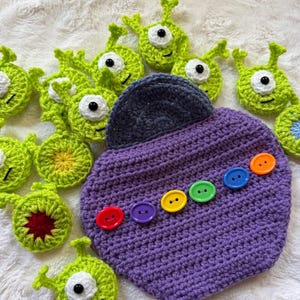 May include: A purple crocheted pouch with a dark grey flap and a row of six colourful buttons, surrounded by several small, round, light green crocheted alien-like figures with single white eyes and antennae on a white textured surface.