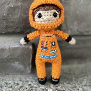 May include: A crocheted doll wearing an orange racing suit with a hood. The suit features the McLaren logo and various sponsor patches including Mastercard, DP World, and Dell Technologies. The doll has brown hair, black eyes, and black cuffs.