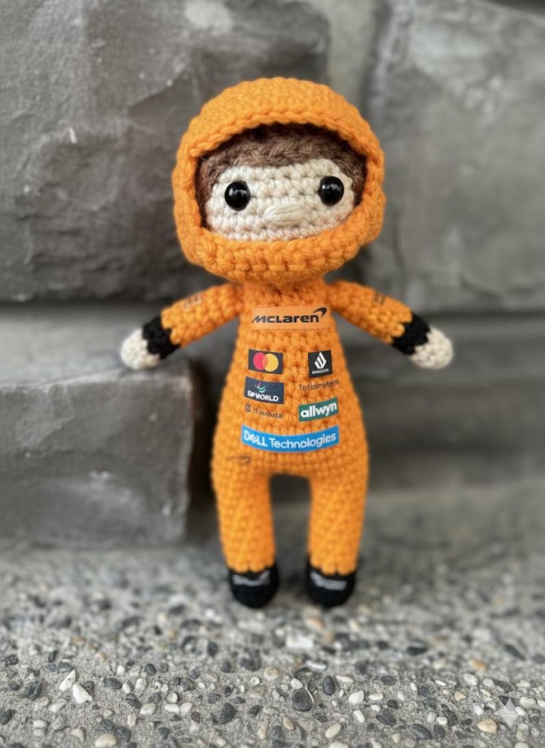 May include: A crocheted doll wearing an orange racing suit with a hood. The suit features the McLaren logo and various sponsor patches including Mastercard, DP World, and Dell Technologies. The doll has brown hair, black eyes, and black cuffs.