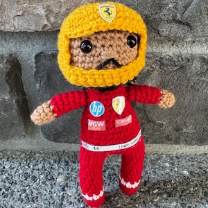 May include: A crocheted doll wearing a yellow racing helmet with a Ferrari logo and a red racing suit. The suit features HP, Ferrari, VGW Play, and Ceva logos, with a white belt displaying the text 44 and LEWIS HAMILTON with a small UK flag.