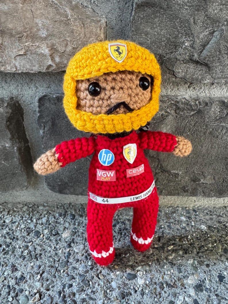 May include: A crocheted doll wearing a yellow racing helmet with a Ferrari logo and a red racing suit. The suit features HP, Ferrari, VGW Play, and Ceva logos, with a white belt displaying the text 44 and LEWIS HAMILTON with a small UK flag.