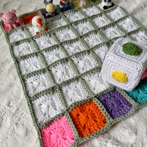 May include: A crocheted blanket featuring a grid of white squares bordered in sage green, with a row of colorful squares at the bottom. Small plastic animal figurines are placed along the top edge, and a white crocheted cube with colored circles sits on the blanket.