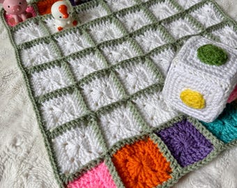May include: A crocheted blanket featuring a grid of white squares bordered in sage green, with a row of colorful squares at the bottom. Small plastic animal figurines are placed along the top edge, and a white crocheted cube with colored circles sits on the blanket.