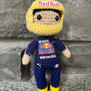 May include: A crochet doll of a race car driver wearing a dark blue racing suit with Red Bull, Mobil 1, Honda, and Puma logos. The doll has a large yellow helmet with a Red Bull logo, black eyes, and tan skin. A small Mexican flag is on the suit.