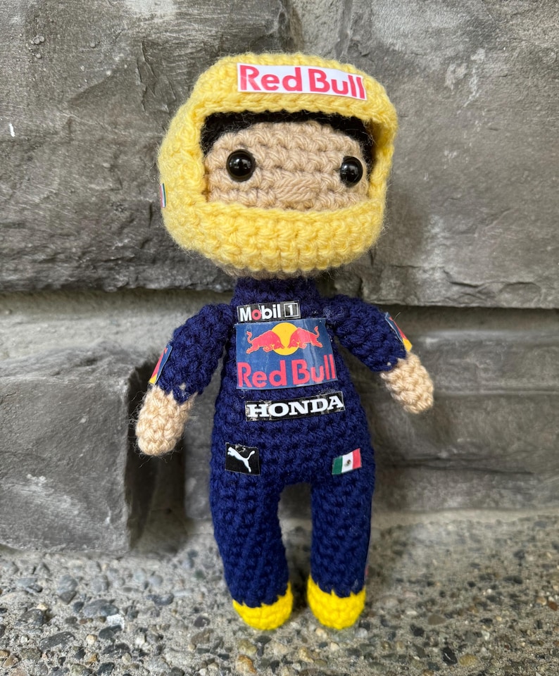 May include: A crochet doll of a race car driver wearing a dark blue racing suit with Red Bull, Mobil 1, Honda, and Puma logos. The doll has a large yellow helmet with a Red Bull logo, black eyes, and tan skin. A small Mexican flag is on the suit.