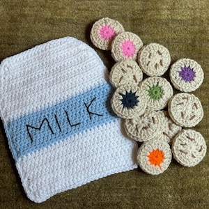 May include: A white and light blue crocheted milk carton shape with the word MILK embroidered in black thread, accompanied by a collection of small, round, beige crocheted cookies featuring centers in pink, purple, green, black, and orange.
