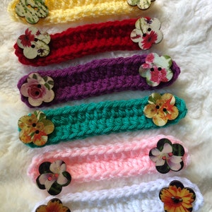 May include: Six crocheted ear savers with floral fabric buttons. The ear savers are in yellow, red, purple, teal, pink, and white.