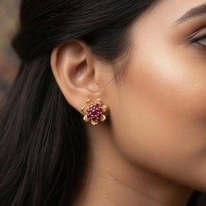 Puede incluir: Pendientes de botón dorados con forma de flor y un grupo de pequeñas piedras redondas de color rubí en el centro. Los pendientes están colocados sobre la piel, mostrando el diseño y el contraste de color. Los pendientes son de diseño floral.